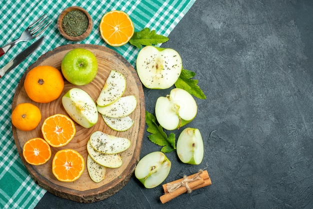 Вид сверху нарезки яблок и мандаринов с порошком сушеной мяты на деревянной доске, вилкой с корицей и ножом на зеленой скатерти на темном фоне