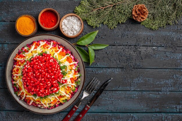 Top view Christmas dish dish with pomegranate carrots potatoes next to the bowls of different spices spruce branches with cone on the table