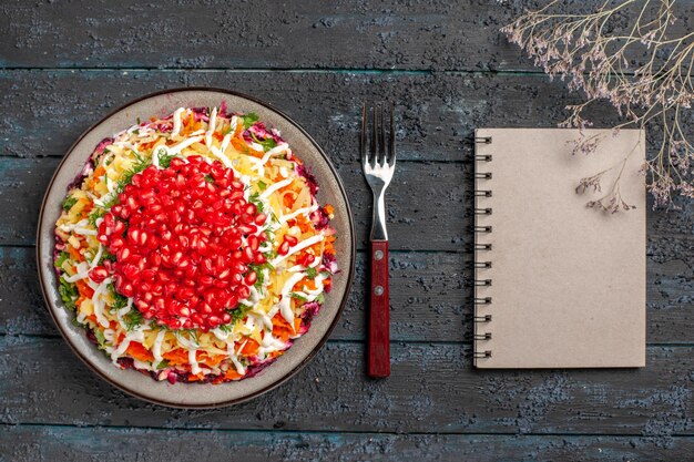 Top view Christmas food tasty Christmas dish with seeds of pomegranate next to the fork tree branches and white notebook on the grey table