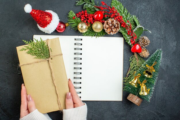 Top view of christmas mood with fir branches santa claus hat hand holding gift box on dark background