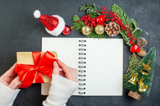 Top view of christmas mood with fir branches santa claus hat hand holding gift box with red ribbon on dark background