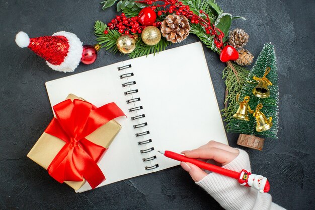 暗い背景の上のノートにペンを持っているモミの枝サンタクロース帽子の手でクリスマス気分の上面図