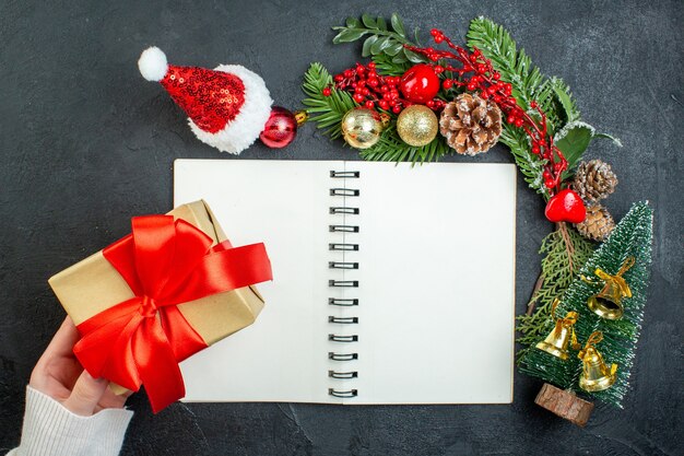 Вид сверху на рождественское настроение с еловыми ветками шляпа санта-клауса рука держит рождественскую елку подарочную коробку с красной лентой на темном фоне