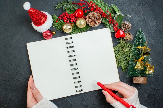 Top view of christmas mood with fir branches xsmas tree santa claus hat hand holding a pen on spiral notebook on dark background