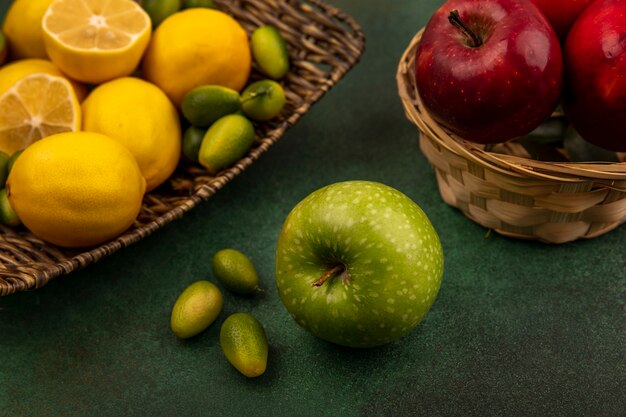 Top view of citrus fruit lemons on a wicker tray with kinkans with green apple on a green surface