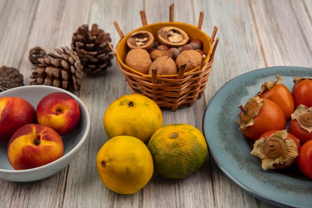 Top view of citrus fruit tangerines with persimmons on a plate with peaches on a bowl with nuts on a bucket on a grey wooden surface