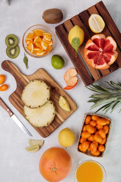 Top view of citrus fruits as cut pineapple on cutting board and cut grapefruit on box with orange kiwi lime with orange juice on white background