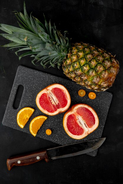Top view of citrus fruits as grapefruit orange pineapple and kumquat with knife on cutting board on black surface