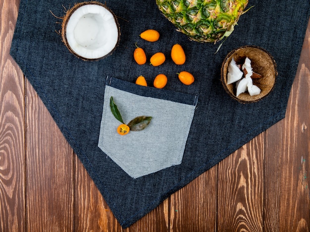 Free photo top view of citrus fruits as half cut coconut with coconut slices in shell kumquats pineapple on jeans cloth and wooden background