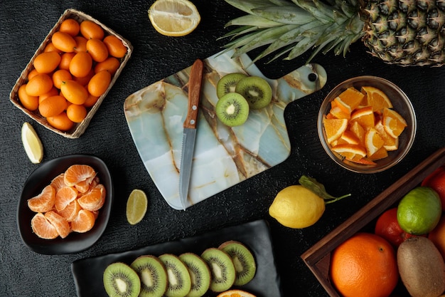 Top view of citrus fruits as kiwi with knife on cutting board tangerine kumquats lemon pineapple and others on black background