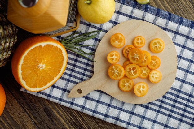 Top view of citrus fruits as kumquat slices on cutting board and lemon cut orange scallion with grater on plaid cloth on wooden background