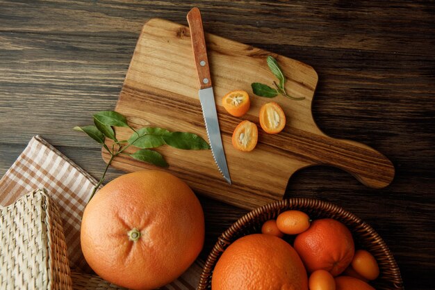 Top view of citrus fruits as kumquat slices with leaves and knife on cutting board with orange tangerine kumquat on wooden background