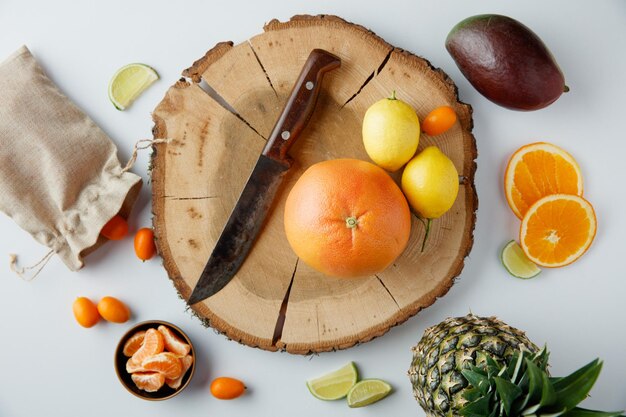 Top view of citrus fruits as lemon kumquat and tangerine with knife on tree stump with avocado pineapple lime and tangerine slices and kumquats spilling out of sack on white background