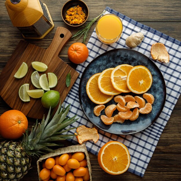 Top view of citrus fruits as orange and tangerine slices in plate with orange juice ginger tangerine shell on plaid cloth with orange zest kumquats lime pineapple with grater on wooden background