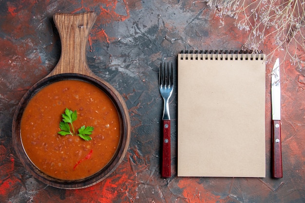 Top view of classic tomato soup on a brown cutting board and notebook on mixed color background