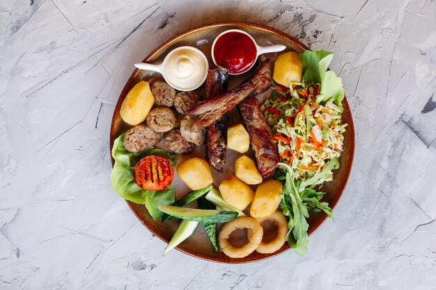 Top view of clay plate full of appetizers for beer including vegetable salad boiled potatoes grilled chicken legs roasted goldy onion and sausages cucmbers tomatoes and sauces ketchup and mayo