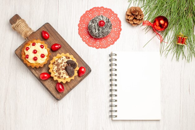 Top view cocoa and berry tarts and cornels on the chopping board cocoa cake and the pine tree leaves with christmas toys and a notebook on the white wooden ground
