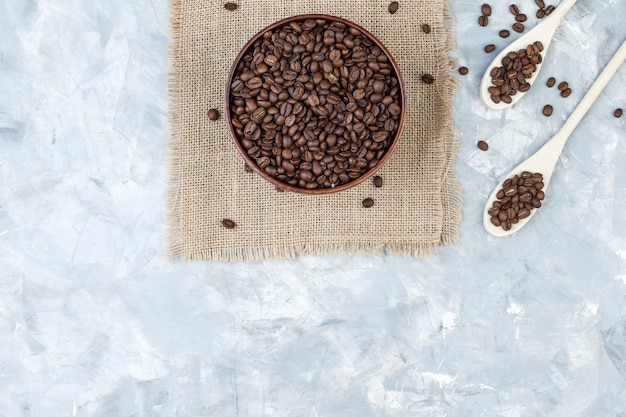 Top view coffee beans in bowl and wooden spoons on plaster and piece of sack background. horizontal