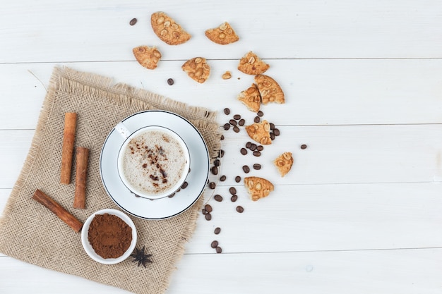 Free photo top view coffee in cup with coffee beans, grinded coffee, cookies, cinnamon sticks on wooden and piece of sack background. horizontal