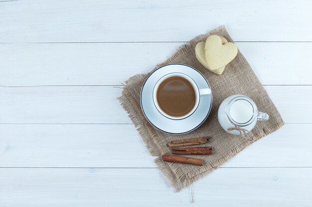 Вид сверху кофе в чашке с печеньем, палочками корицы, молоком на деревянном и куске фона мешка.