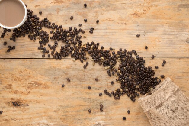 Top view of coffee cup with roasted and raw coffee beans falling from sack on table