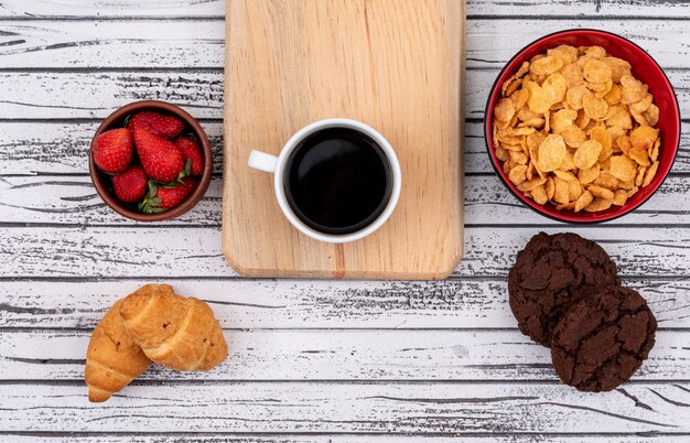 Top view of coffee with cookies and croissants, cornflakes with strawberry on white wooden surface horizontal