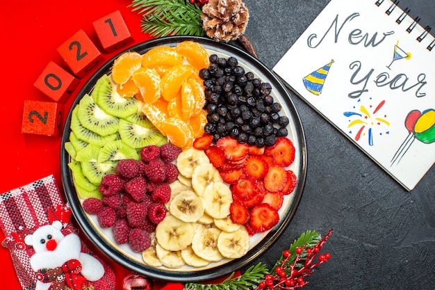 Top view of collection of fresh fruits on dinner plate decoration accessories fir branches and numbers christmas sock on a red napkin next notebook with new year drawings on a black background