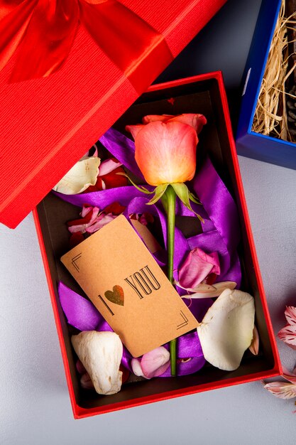 Top view of color color rose flower with purple ribbon and small brown paper card in a red present box on white table