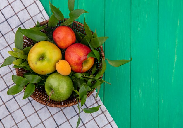 Вид сверху цветных яблок в корзине с ветвями листьев с белым клетчатым полотенцем на зеленой поверхности