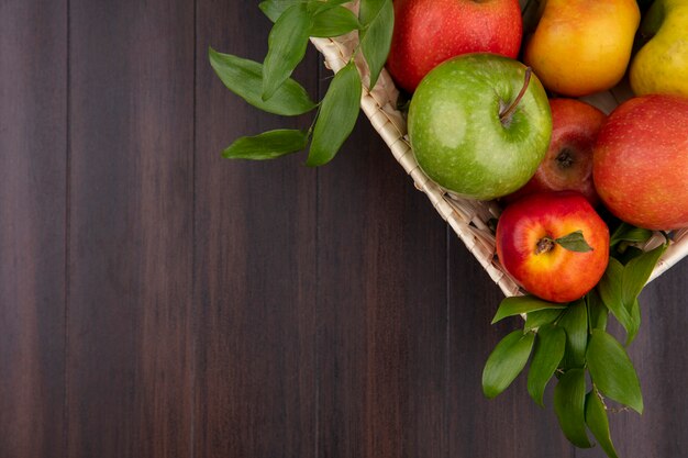 Вид сверху цветных яблок в корзине с ветвями листьев на деревянной поверхности