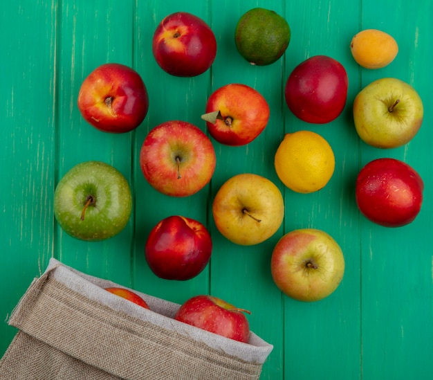 Top view of colored apples with peaches lemon and lime in a burlap bag on a green surface