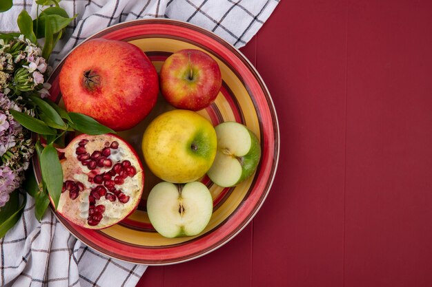 Top view of colored apples with pomegranate on a plate with a checkered white towel on a red surface