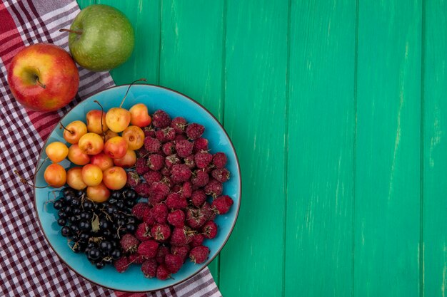 Top view of colored apples with raspberries white cherries and black currants on a plate on a green surface