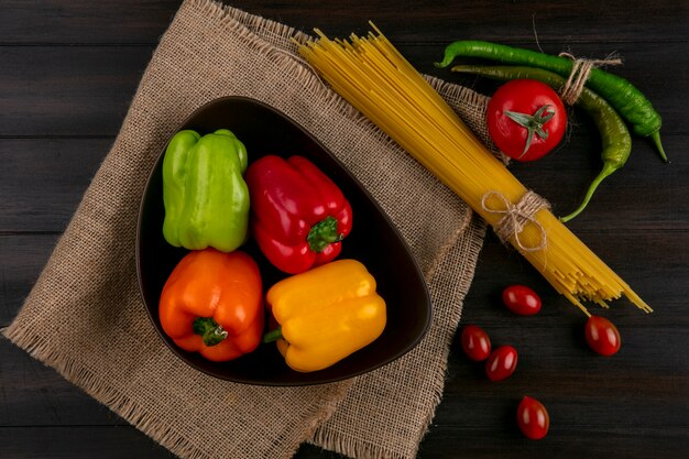 Top view of colored bell peppers in a bowl with raw spaghetti chili peppers and tomatoes on a wooden surface