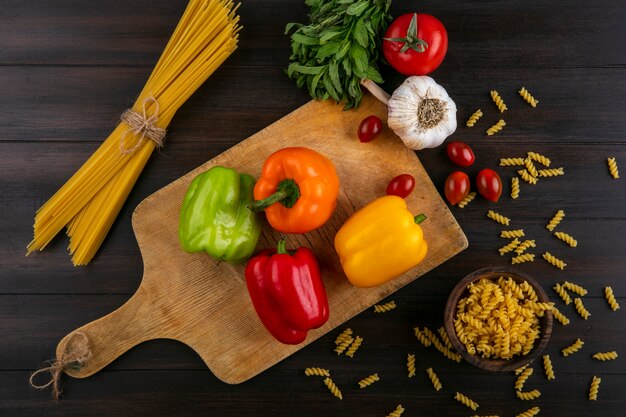 Top view of colored bell peppers on a cutting board with garlic washed and raw spaghetti on a wooden surface