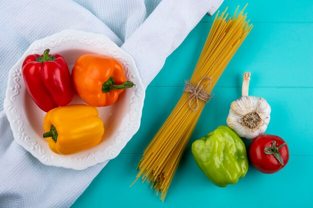 Top view of colored bell peppers in a white plate on a white towel with raw spaghetti tomato and garlic