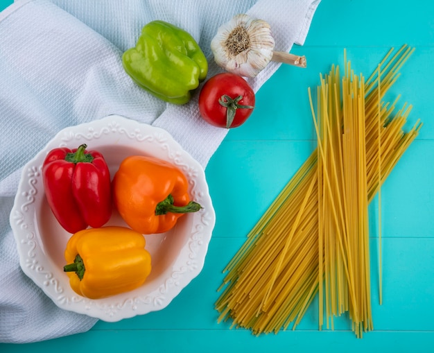 Top view of colored bell peppers in a white plate on a white towel with raw spaghetti tomato and garlic