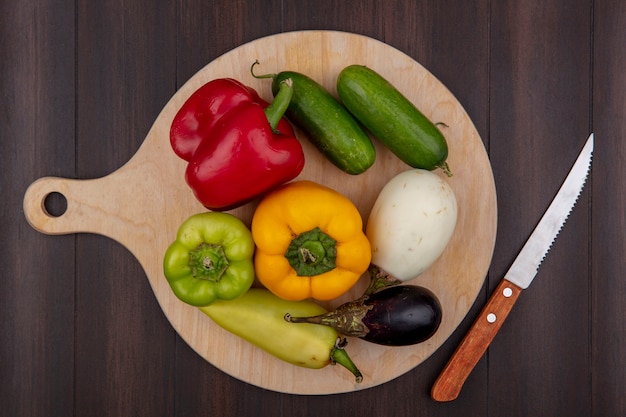 Top view colored bell peppers with bklazana and cucumber on a cutting board with a knife  on a wooden background