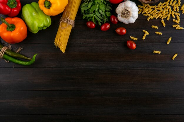 Top view of colored bell peppers with raw spaghetti and pasta garlic and tomatoes on a wooden surface