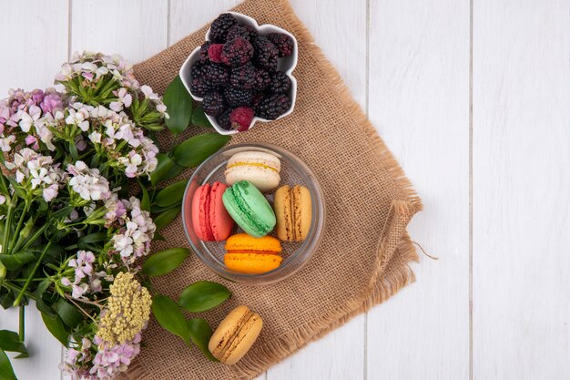 Top view of colored macarons in a jar with flowers and blackberries on a white surface