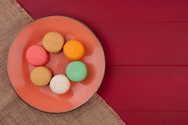 Top view of colored macarons on an orange plate on a beige napkin on a red surface