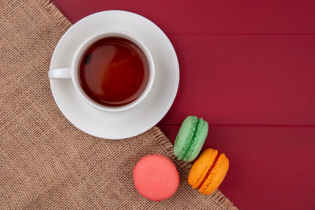 Top view of colored macarons with a cup of tea on a beige napkin on a red surface