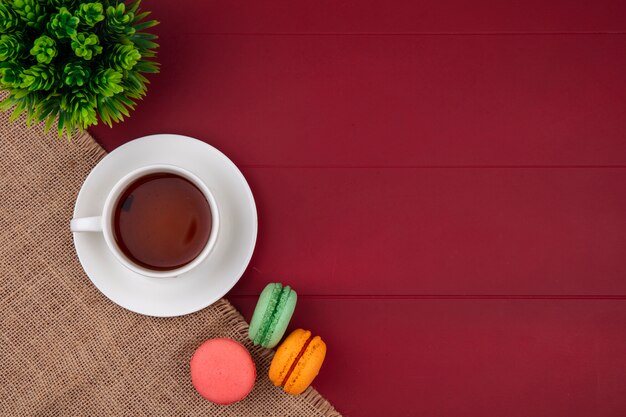 Top view of colored macarons with a cup of tea on a beige napkin on a red surface