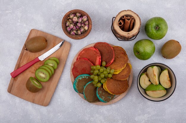 緑のリンゴと白い背景の上のシナモンとボード上のナイフとキウイのスタンドに緑のブドウと色のパンケーキの上面図