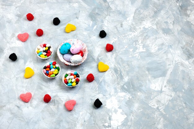 Вид сверху красочные конфеты внутри маленьких тарелок вместе с мармеладными конфетами в форме сердца на серо-белом фоне.