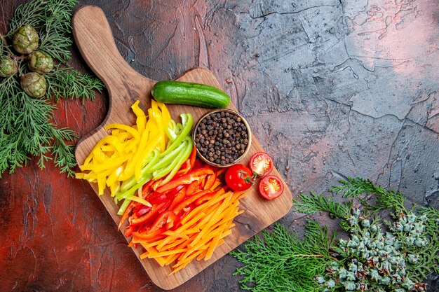 Top view colorful cut peppers black pepper tomatoes cucumber on cutting board pine branches on dark red table free space