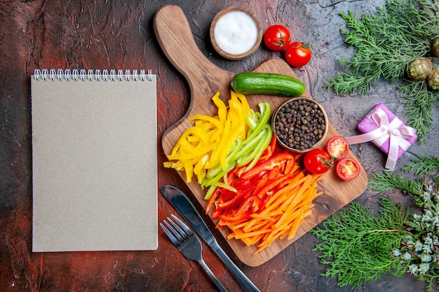 Top view colorful cut peppers black pepper tomatoes cucumber on cutting board salt fork knife notebook on dark red table