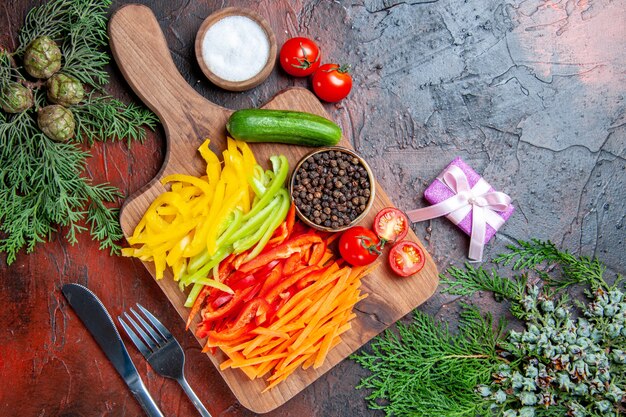 Top view colorful cut peppers black pepper tomatoes cucumber on cutting board salt fork and knife small gift on dark red table