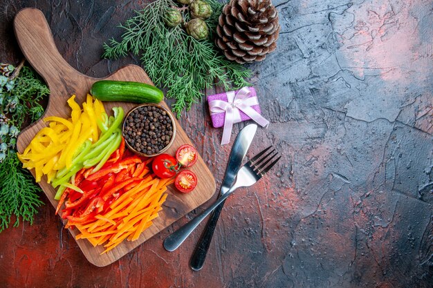 Top view colorful cut peppers black pepper tomatoes cucumber on cutting board small gift fork and knife pine branches on dark red table free space