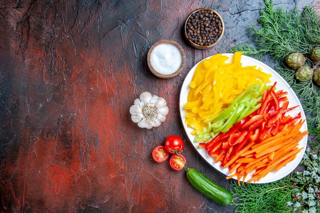 Top view colorful cut peppers on plate salt and black pepper tomatoes garlic cucumber on dark red table free space